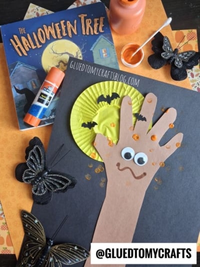 Halloween craft scene with cut-out paper hand on black paper, googly eyes, orange dots, bats, glue stick, orange paint, butterflies, and The Halloween Tree book—perfect for fall keepsake ideas. The @GLUEDTOMYCRAFTS label is at the bottom.