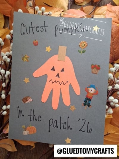 A gray paper with Cutest pumpkin in the patch 26 written on it, decorated with fall stickers and an orange handprint shaped like a jack-o-lantern. Autumn leaves and stars surround the artwork—a charming example of fall keepsake ideas to treasure seasonal memories.
