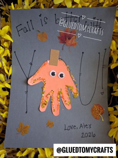 A gray card with Fall is better with you! written on it. An orange handprint, decorated with googly eyes and glitter, is clipped to the card, surrounded by fall leaves. Looking for fall keepsake ideas? This sweet creation is finished with Love, Alex 2026 at the bottom.