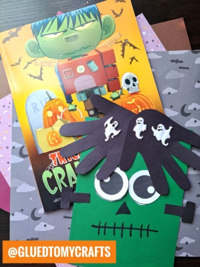 A handprint Frankenstein craft made from green paper, googly eyes, and a black handprint cutout for hair with ghost stickers is displayed on top of Halloween-themed papers and a Frankenstein book.