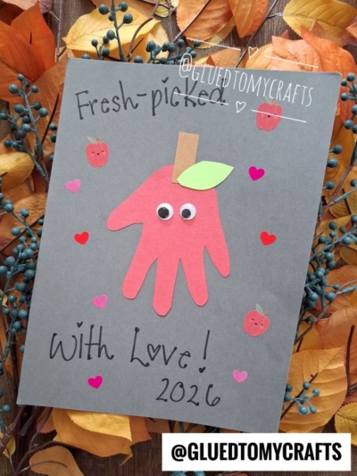 A handmade card features a red handprint apple with googly eyes on gray paper, surrounded by small apples and hearts. Perfect for those seeking fall keepsake ideas, the text reads Fresh picked with love! 2026. Autumn leaves and berries border the scene.