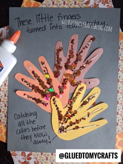 Four handprint cutouts decorated with glitter and sequins are glued on gray paper, making for one of the sweetest fall keepsake ideas. A bottle of glue sits to the side. Text on the paper reads: “These little fingers turned into leaves today.”.
