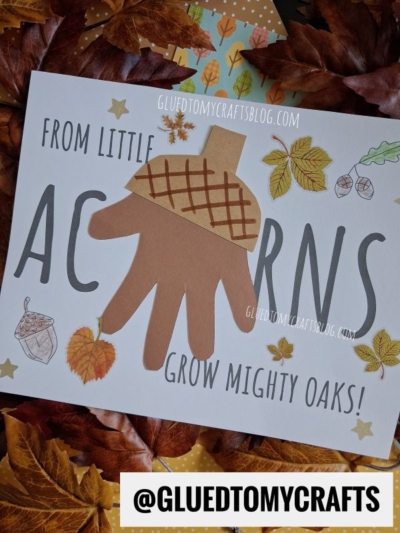 A craft sheet says “From little acorns grow mighty oaks!” with leaves and acorns. A brown handprint cutout and paper acorn are glued as decoration, making this one of the sweetest fall keepsake ideas. Dried leaves and a paper with @GLUEDTOMYCRAFTS surround the sheet.