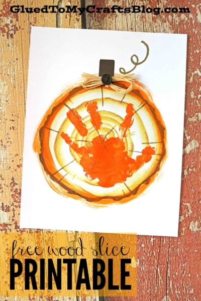 A children’s craft featuring an orange handprint on a wood slice printable, decorated as a pumpkin keepsake, clipped to a rustic wood background. Text reads free wood slice PRINTABLE.