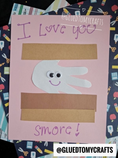 A handmade card with "I Love You Smore!" written in purple marker, a white paper hand with googly eyes and a smile, and brown construction paper strips resembling a s'more—perfect for fall keepsake ideas.