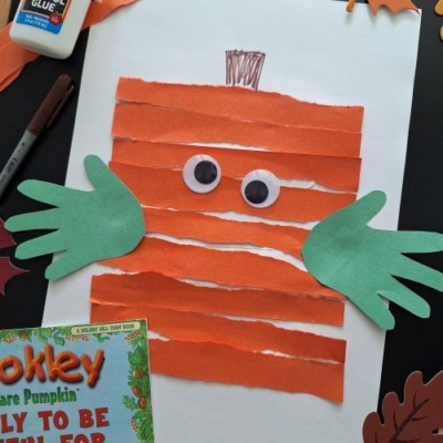 A fun fall keepsake idea: a kids’ pumpkin craft made with torn orange paper, green handprint cutouts for leaves, large googly eyes, and a drawn stem, surrounded by glue, paper leaves, and a pumpkin book.