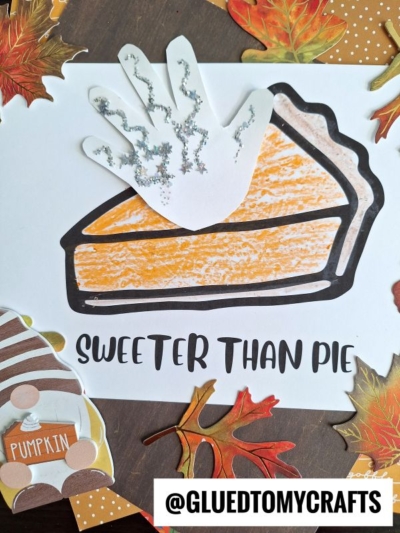 A handprint decorated with glitter appears as whipped cream on a colored-in pumpkin pie drawing—an adorable option for fall keepsake ideas. Text below reads SWEETER THAN PIE. Fall leaves and crafting materials surround the artwork.