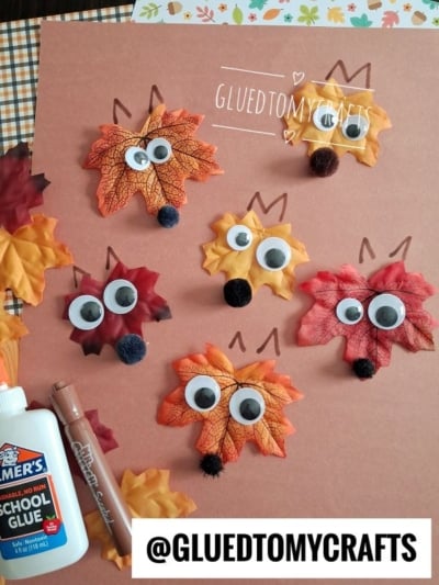 Five fox faces made from fake autumn leaves, googly eyes, and small black pom-poms are arranged on brown paper—perfect inspiration if you’re searching for leaf kid craft ideas. A glue bottle and brown marker are nearby. @GLUEDTOMYCRAFTS is written at the bottom.