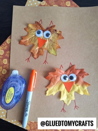 Two owl crafts made from artificial autumn leaves, googly eyes, and orange paper beaks are glued to brown paper. A tape runner, orange Sharpie, and patterned paper are nearby—fun leaf kid craft ideas! @GLUETOMYCRAFTS is labeled at the bottom.