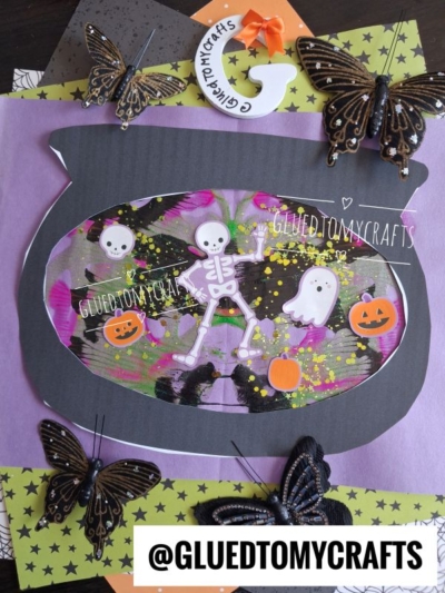 A Halloween-themed paper craft featuring a fishbowl with skeletons, ghosts, pumpkins, and colorful paint splat effects, surrounded by artificial butterflies. A whimsical witch’s cauldron adds to the spooky charm. @GLUEDTOMYCRAFTS is displayed at the bottom.