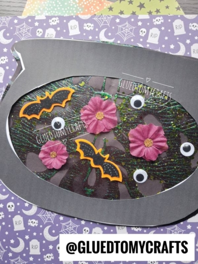 A Halloween-themed craft featuring a gray fishbowl shape reminiscent of a paint splat witch’s cauldron, adorned with pink flowers, bats, googly eyes, and sparkly green swirls on a purple background with stars, moons, and spiderweb patterns.