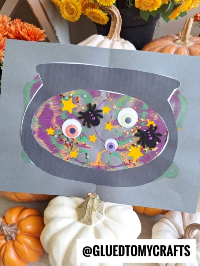 A colorful paint splat witch’s cauldron paper craft, decorated with googly eyes, star stickers, spiders, and glitter. Pumpkins and flowers are in the background. The text @GLUEDTOMYCRAFTS is at the bottom.