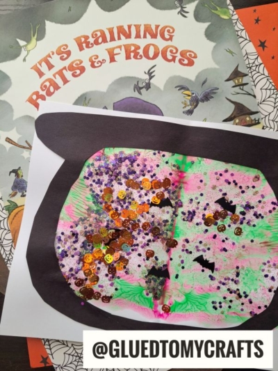 A colorful paint splat witch's cauldron craft with glitter, sequins, and bat shapes sits atop the Halloween-themed book titled Its Raining Bats & Frogs. The handle @GLUEDTOMYCRAFTS appears at the bottom.