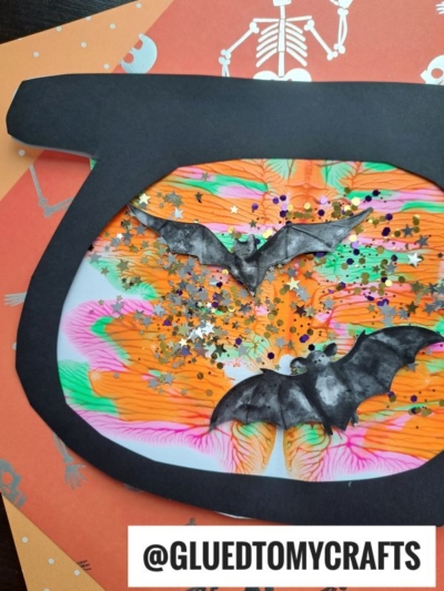 A Halloween craft featuring a black cat-shaped frame with colorful swirled paper, shiny star confetti, and two black bat cutouts inside—plus hints of paint splat for a magical touch. Orange paper with skeletons is in the background.