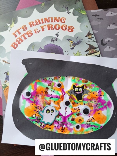 A colorful Halloween-themed paint splat witch's cauldron craft with googly eyes, bat and ghost stickers, and swirls of neon paint sits on top of an Its Raining Bats & Frogs book and patterned papers. @GLUEDTOMYCRAFTS.