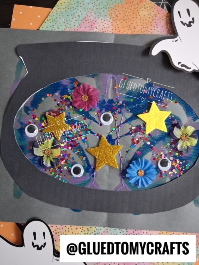 A colorful craft featuring a decorated paint splat witch’s cauldron with googly eyes, flowers, star shapes, and glitter, surrounded by smiling ghost cutouts. The handle @GLUEDTOMYCRAFTS appears at the bottom.