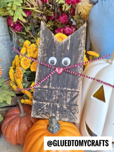 A charming paper bag black cat craft with googly eyes, a red heart nose, and sparkly pink pipe cleaner whiskers, displayed among pumpkins, flowers, and a white jack-o-lantern for a festive touch.