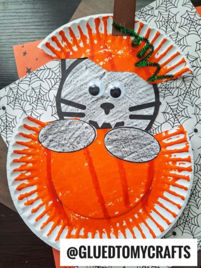 Paper plate craft featuring a black cat in pumpkin, peeking over an orange pumpkin with googly eyes, spiderweb-patterned background, and @GLUEDTOMYCRAFTS watermark at the bottom.