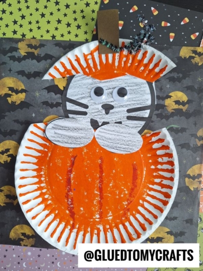 A Halloween craft features a black cat in pumpkin made from paper plates, painted orange and white, with googly eyes, pipe cleaner whiskers, and a brown stem, resembling a pumpkin, on a festive Halloween-themed background.