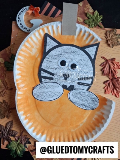 Paper plate painted orange to look like a pumpkin, with a gray paper cat face, paws, and googly eyes attached—creating the whimsical look of a black cat in pumpkin. Displayed on a wood surface with autumn leaves and finished with a brown craft stick for a tail. @GLUEDTOMYCRAFTS text at the bottom.