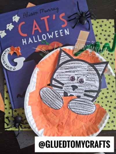 A childrens book titled Cats Halloween, a spider toy, and a paper plate craft of a gray cat with googly eyes on orange tissue paper are featured, along with a charming black cat in pumpkin to add festive flair. The handle @gluedtomycrafts is displayed at the bottom.