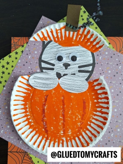 Paper plate craft of a black cat in pumpkin, with googly eyes, black paper whiskers, and colored paper details. The adorable cat holds an orange pumpkin and sits on patterned Halloween-themed paper.