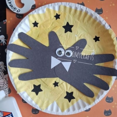 A Halloween craft with black paper bat wings shaped like hands on a yellow circle, surrounded by stars, with a bow tie in the center. There’s school glue and a decorated orange paper in the background—perfect inspiration if you’re looking for creative fall keepsake ideas.