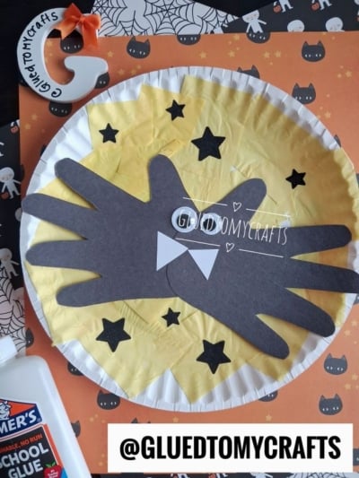 A Halloween craft with black paper bat wings shaped like hands on a yellow circle, surrounded by stars, with a bow tie in the center. There’s school glue and a decorated orange paper in the background—perfect inspiration if you’re looking for creative fall keepsake ideas.