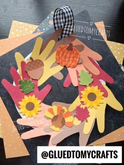 A fall wreath made from paper handprints, decorated with pumpkins, sunflowers, leaves, an acorn, and a turkey, plus a black-and-white checkered ribbon—one of the cutest fall keepsake ideas for kids!.