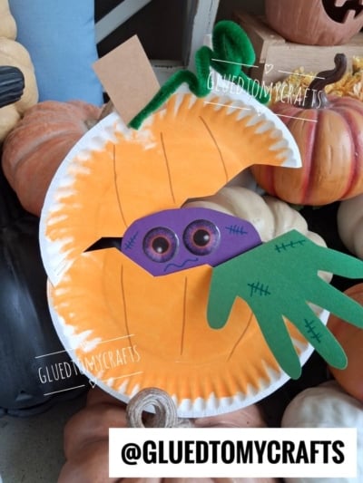 Paper pumpkin craft with a playful pumpkin monster peeking out, featuring big eyes and a green felt hand, surrounded by real pumpkins. GLUEDTOMYCRAFTS is watermarked on the image, with the handle at the bottom.