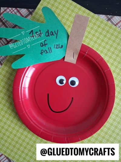 A red paper plate apple craft with googly eyes, a smiling face, a green handprint leaf, and a brown paper stem sits on a yellow plaid background. Text on the leaf reads, “1st day of fall 126.” This adorable project is perfect for those looking for creative fall keepsake ideas.