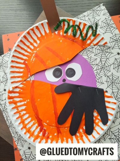 A Halloween craft featuring a pumpkin monster made from an orange painted paper plate with purple construction paper eyes, a black paper hand mouth, a brown stem, and a green pipe cleaner vine. Spiderweb-patterned paper is underneath.