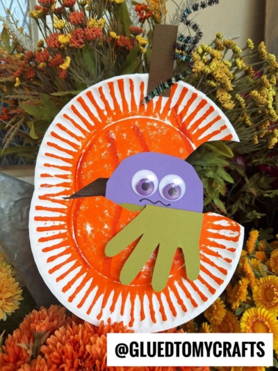 A pumpkin monster craft made from a painted orange paper plate, with a purple and green paper monster face, googly eyes, and a green handprint, set amid autumn flowers. @GLUEDTOMYCRAFTS text is visible.