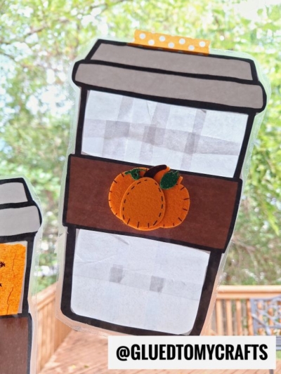 Paper coffee cup craft with a brown sleeve featuring an orange pumpkin, creating a charming stained glass pumpkin spice latte effect when sunlight streams through. The cup is taped to a window, with green foliage and a wooden railing visible outside. Text at the bottom reads @GLUEDTOMYCRAFTS.