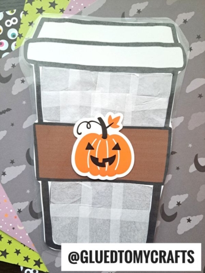 A paper craft of a to-go coffee cup decorated with a jack-o-lantern, reminiscent of a stained glass pumpkin spice latte, set on a Halloween-themed background with bats, ghosts, and googly eyes. Text at the bottom reads @GLUEDTOMYCRAFTS.