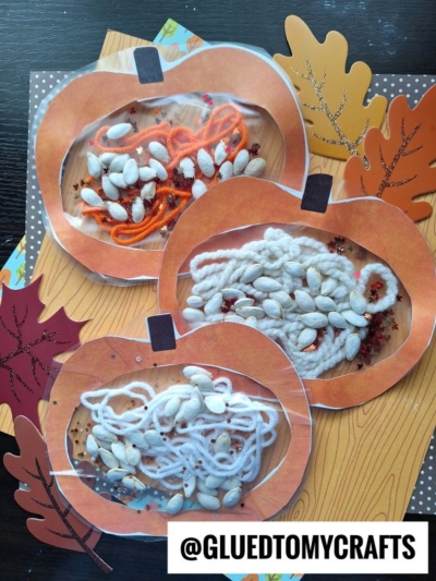 Three paper pumpkins with clear windows reveal what's inside my pumpkin craft—white yarn, orange yarn, white pumpkin seeds, and glitter. Paper leaves surround them on a wood-patterned surface. @GLUETOMYCRAFTS is at the bottom.