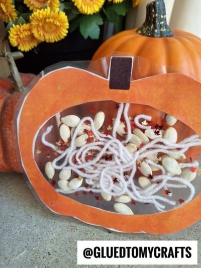 A pumpkin-shaped sensory bag with white yarn, pumpkin seeds, and glitter inside my pumpkin craft, displayed next to artificial pumpkins and yellow flowers. The handle reads @GLUEDTOMYCRAFTS.