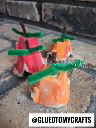 Three painted egg carton pieces resembling apples and pumpkins with green pipe cleaner stems sit on brick, creating a charming egg carton pumpkin patch. The handle @GLUEDTOMYCRAFTS is overlaid on the image.