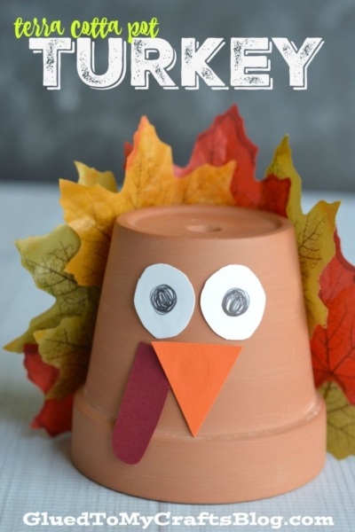 A small terra cotta pot decorated as a turkey with paper eyes, beak, wattle, and colorful fake leaves for feathers—perfect for those who love creative terra cotta pot crafts. Terra Cotta Pot TURKEY and GluedToMyCraftsBlog.com are written at the top and bottom.