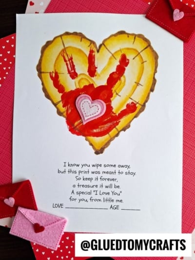 A sheet with a heart-shaped tree trunk illustration, a child’s painted red handprint—capturing their handprint little forever—and a felt heart on top. Below is a poem about preserving a child's touch, surrounded by red and pink paper and felt envelopes.