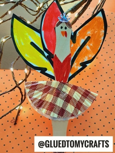 A colorful craft stick turkey ballerina made from a wooden spoon, painted paper for feathers, and a red-and-white checkered paper skirt, with a small blue crown, on an orange polka-dot background. @GLUEDTOMYCRAFTS is at the bottom.