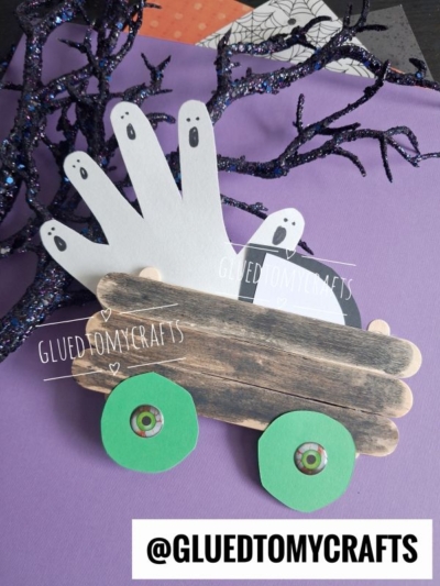 A spooky craft featuring paper ghosts made from white handprints riding in a brown Popsicle Stick Halloween Truck with green wheels, set on a purple surface near a sparkly black branch.