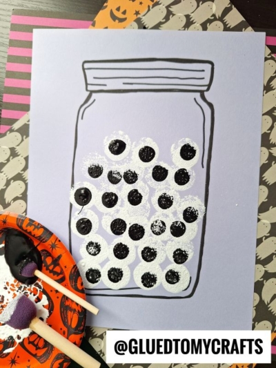 A drawing of a stamped eyeball jar on lavender paper is decorated with clusters of black and white dots resembling googly eyes, next to paint supplies and Halloween-themed paper. Text at the bottom reads @GLUEDTOMYCRAFTS.