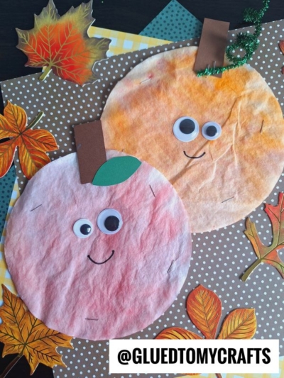 Two round fall crafts, including a coffee filter apple and a pumpkin, are made from dyed coffee filters with googly eyes, paper stems, and smiles. They rest on a polka-dot background decorated with paper autumn leaves. @GLUEDTOMYCRAFTS.