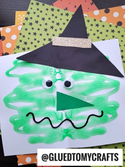 A paint splat witch face is formed by a green paint blot shaped like a witch’s face, with googly eyes, a green triangle nose, a black squiggly mouth, and a black hat on white paper. The background includes orange and green star-patterned papers.