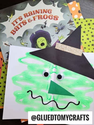 A children’s book titled “It’s Raining Bats & Frogs” with a green witch on the cover sits behind a kid-made paint splat witch face craft, featuring a hat, googly eyes, and wavy mouth. @GLUEDTOMYCRAFTS is written below.