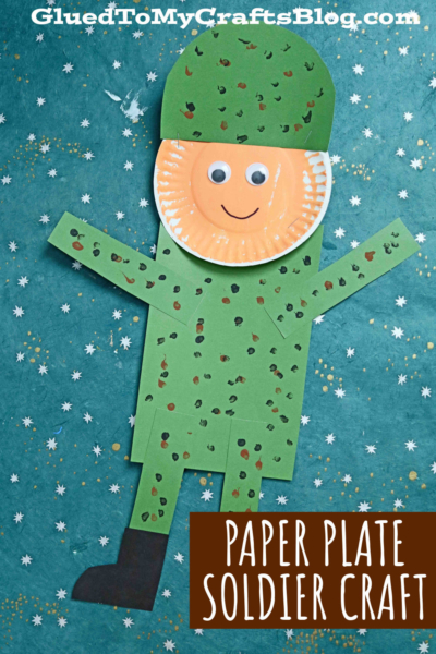 A paper plate soldier craft for kids with a smiling face, green helmet, and dotted uniform, displayed on a blue star-patterned background. Text reads Paper Plate Soldier Craft.