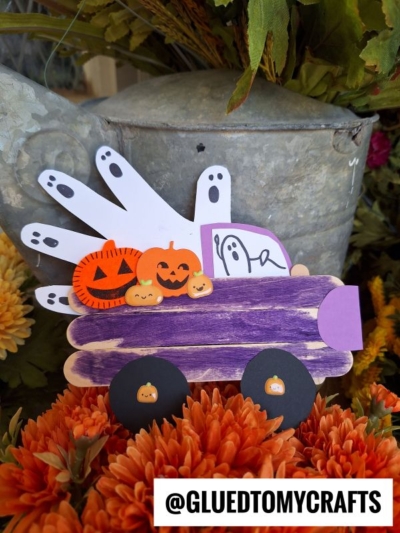 A Halloween craft featuring a Popsicle Stick Halloween Truck painted purple, decorated with paper pumpkins and a handprint ghost, surrounded by orange flowers. @GLUEDTOMYCRAFTS text is at the bottom.