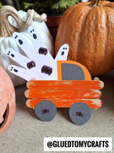 A Popsicle Stick Halloween Truck craft featuring an orange popsicle stick ghost truck with paper details, four white handprint ghosts, bat stickers, and pumpkins. @GLUEDTOMYCRAFTS appears at the bottom.