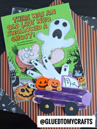 A children’s book titled There Was an Old Lady Who Swallowed a Ghost! is next to a Popsicle Stick Halloween Truck craft, featuring a purple truck carrying pumpkins and ghost cutouts on a striped orange and black background.